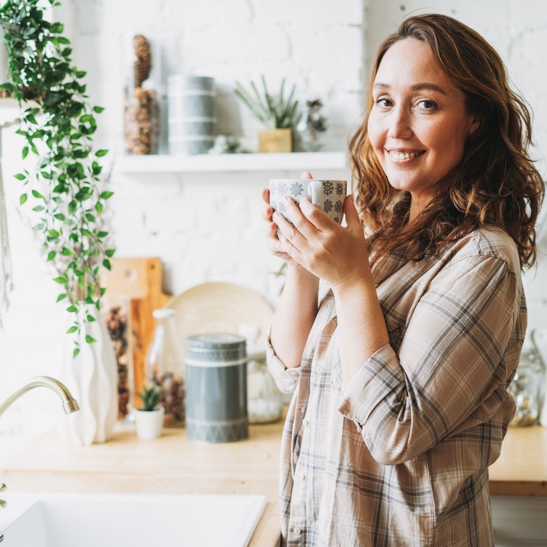 Smiling woman in a plaid shirt holding a patterned mug in a bright kitchen with plants