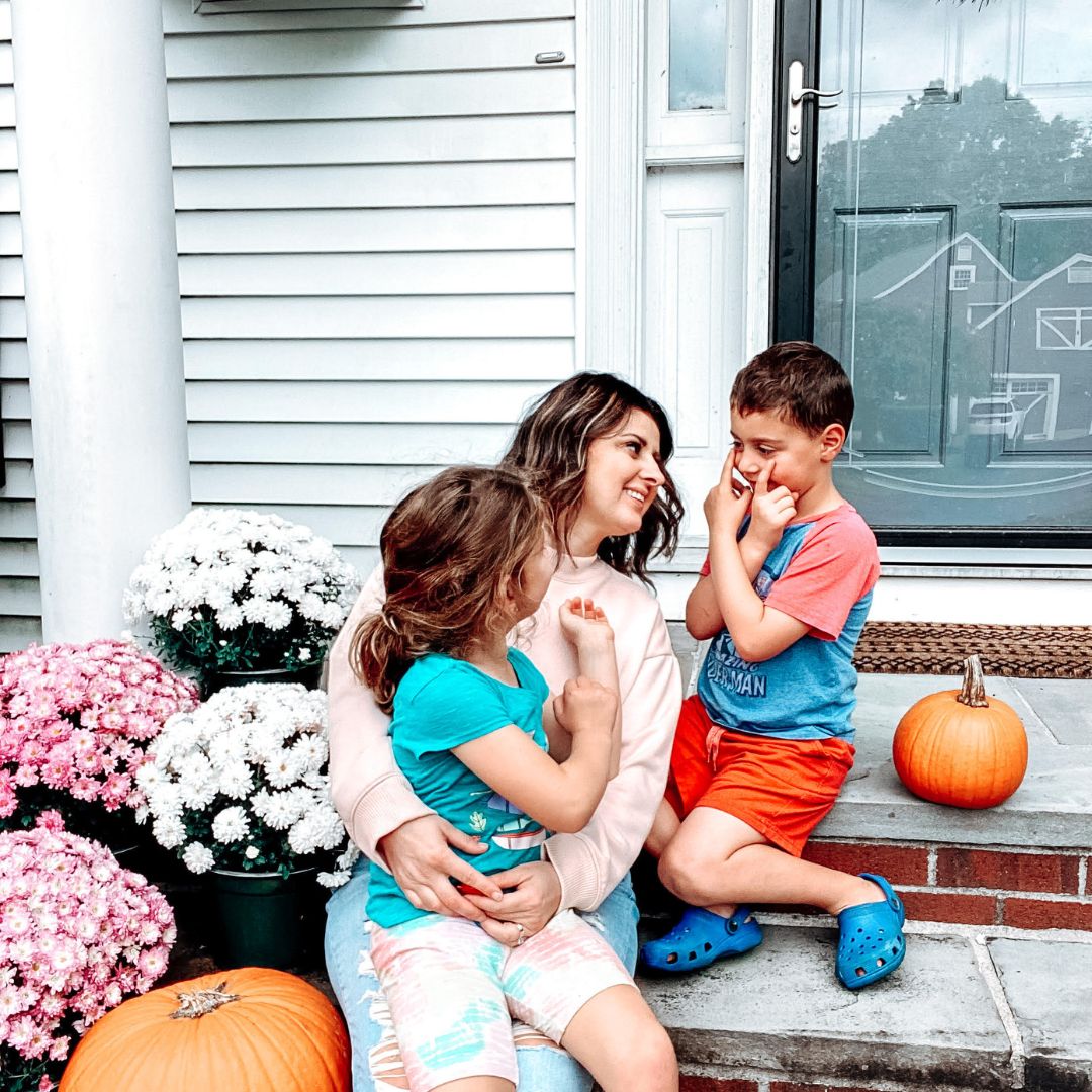 Mother sitting on front steps hugging her daughter while smiling at her son beside pumpkins and potted mums