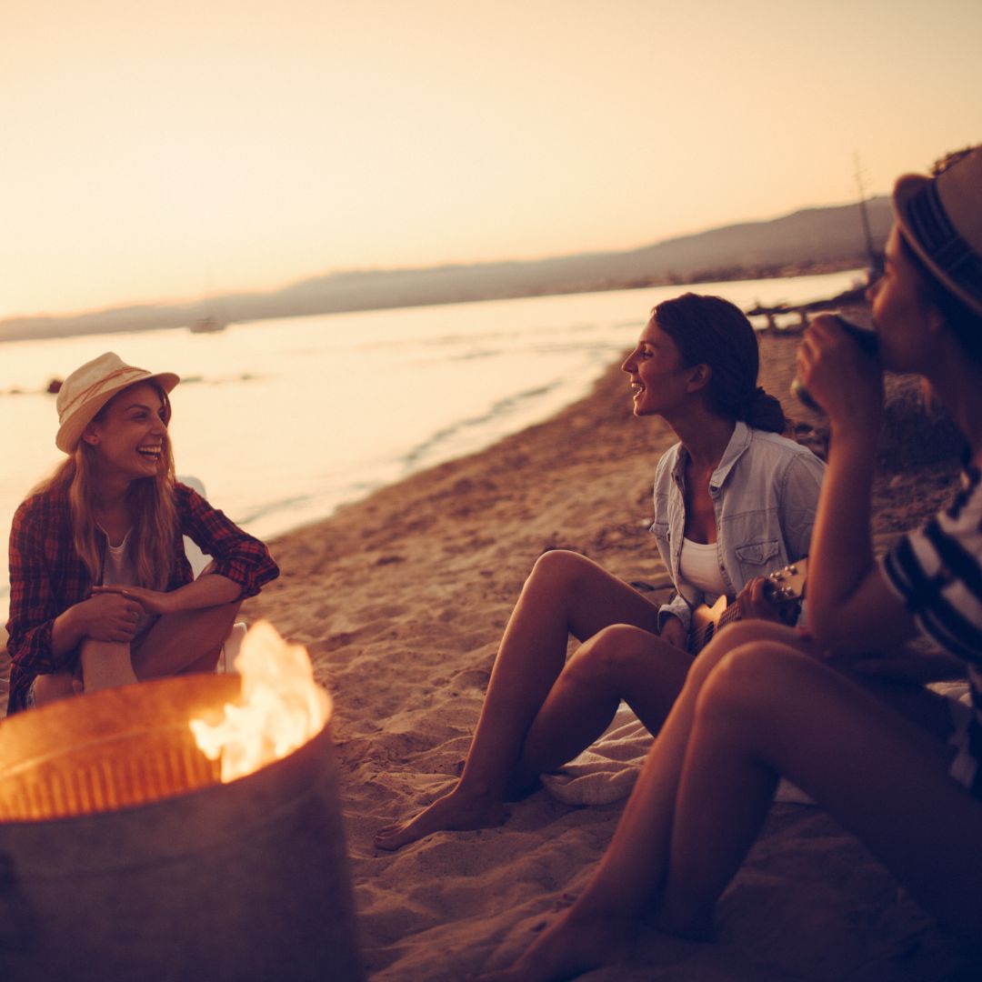 Three friends sitting on a sandy beach at sunset, laughing and talking around a small bonfire
