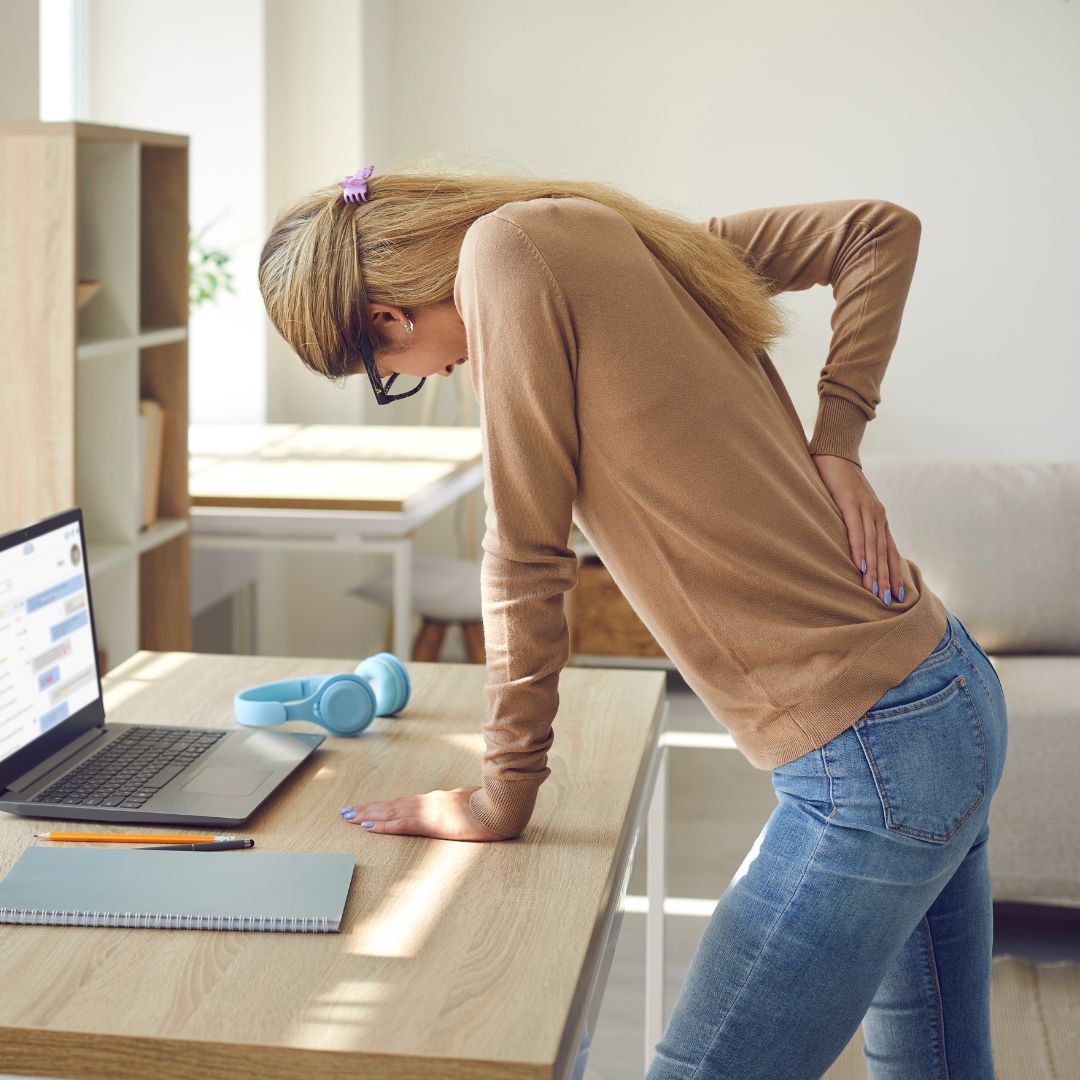 Woman leaning on a desk holding her lower back in a home office, wearing a brown sweater and jeans