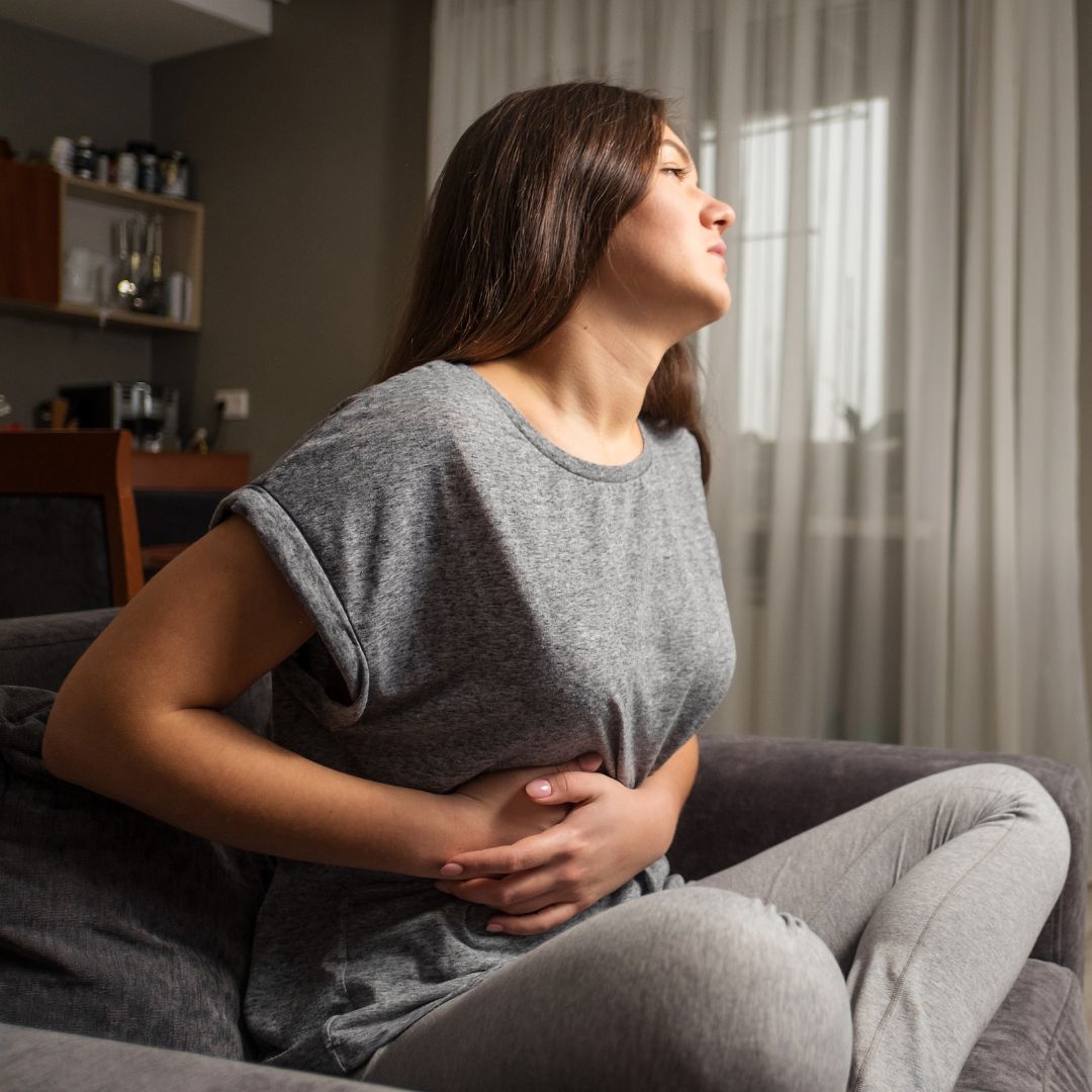 Woman clutching her lower abdomen on a couch, appearing to have menstrual cramps in a living room