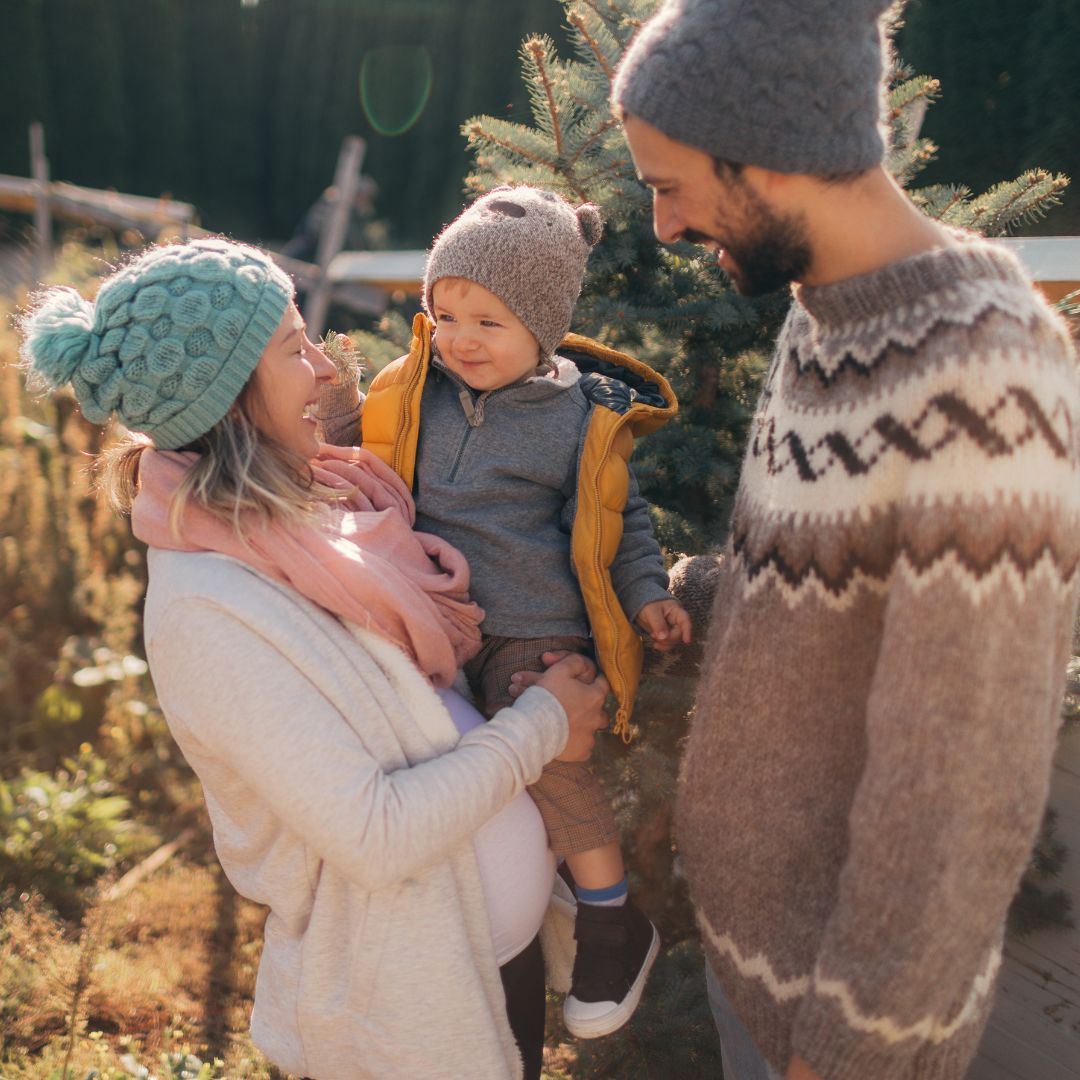 Parents holding a toddler near an evergreen, all wearing winter hats and sweaters in warm sunlight