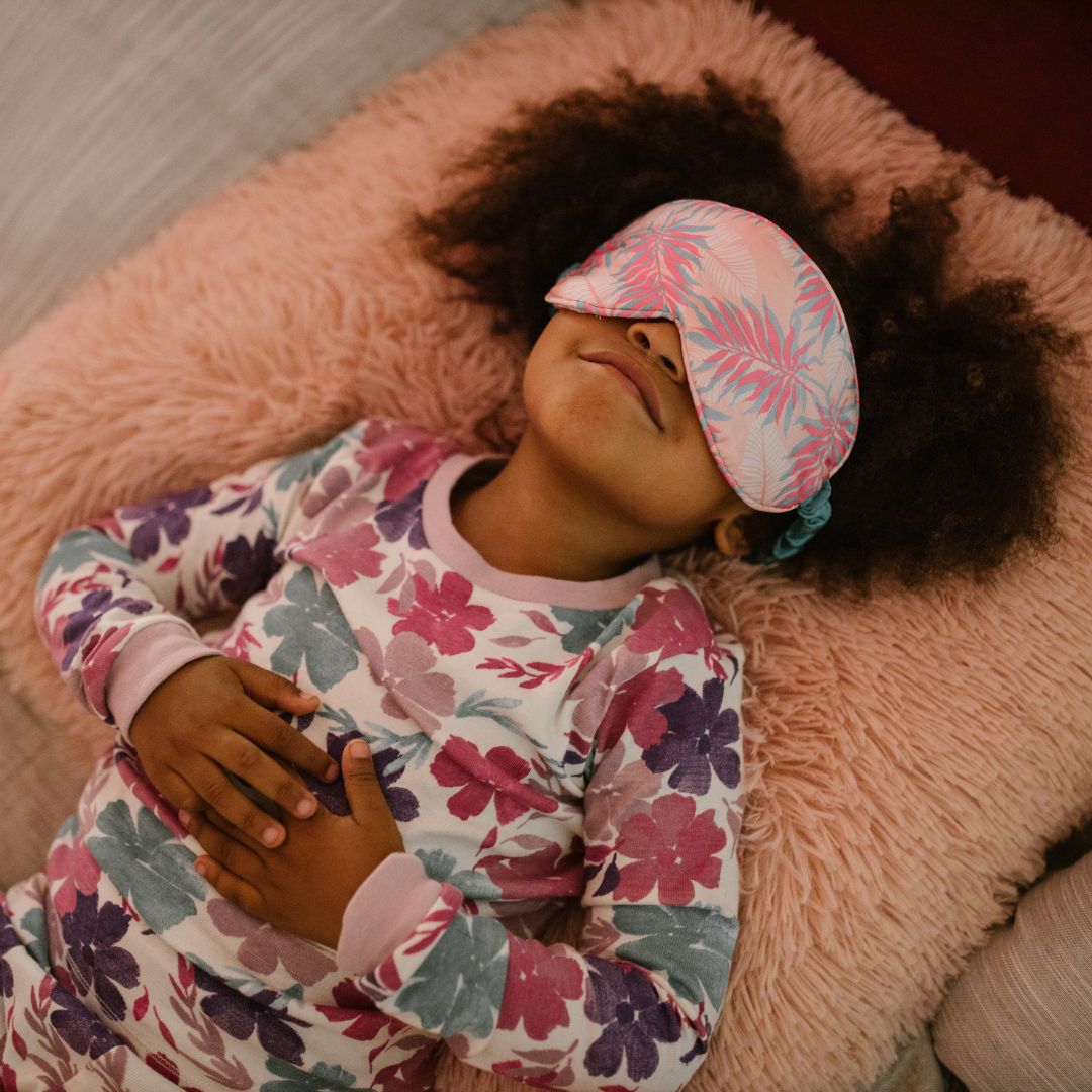 Child lying on a pink fuzzy pillow wearing floral pajamas and a pink leafy sleep mask, resting with hands on stomach