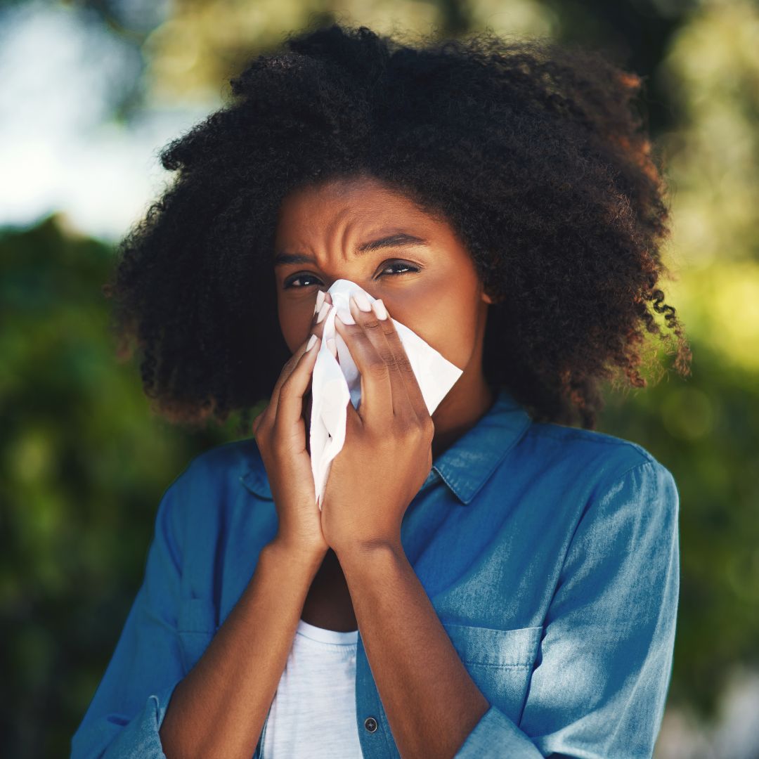 Woman sneezing into a tissue outdoors with a blurred green background