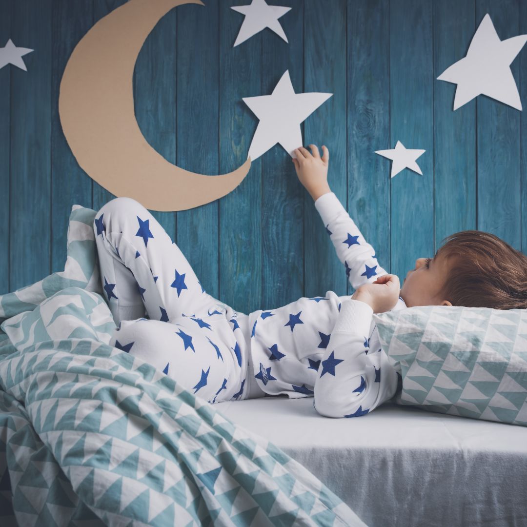 Child lying on bed reaching for paper star on blue wooden wall, wearing white pajamas with blue stars and geometric bedding