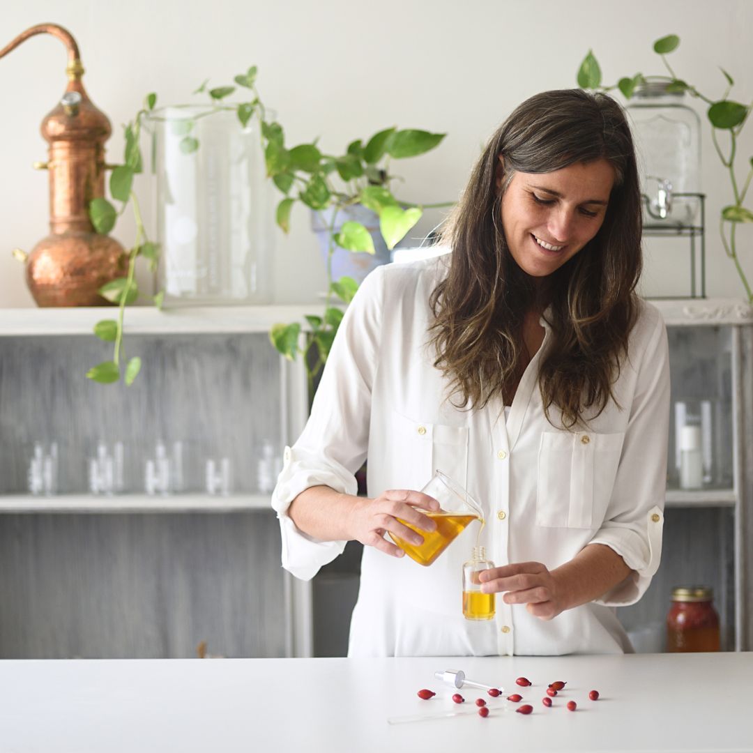 Woman pouring rosehip seed oil into a small dropper bottle on a white counter with scattered rosehip seeds and apothecary jars in the background