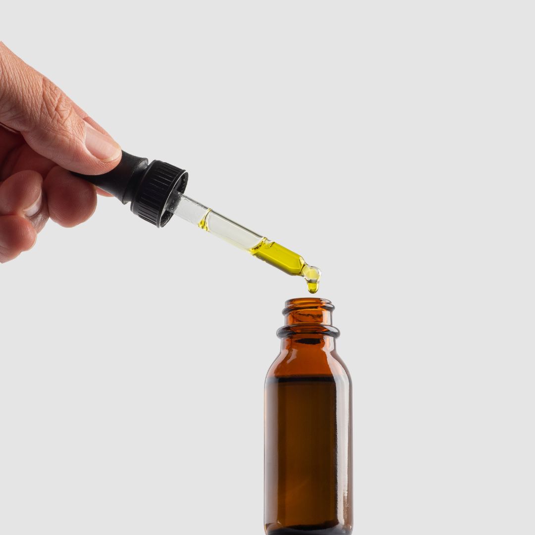 Angled close-up of amber dropper bottle showing a glass dropper releasing a yellow oil droplet on a light gray background
