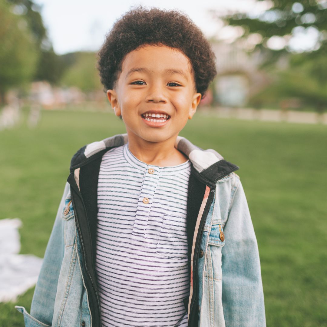 Child smiling while standing in a park wearing a striped shirt and denim jacket