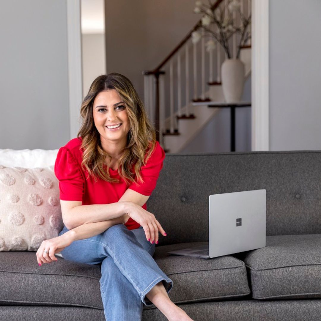 Woman smiling on a gray sofa with a laptop, wearing a red top and jeans in a living room with a staircase