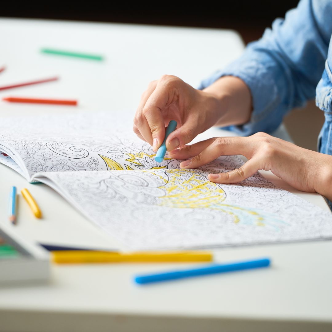 Hands coloring an intricate adult coloring book with colored pencils on a white table