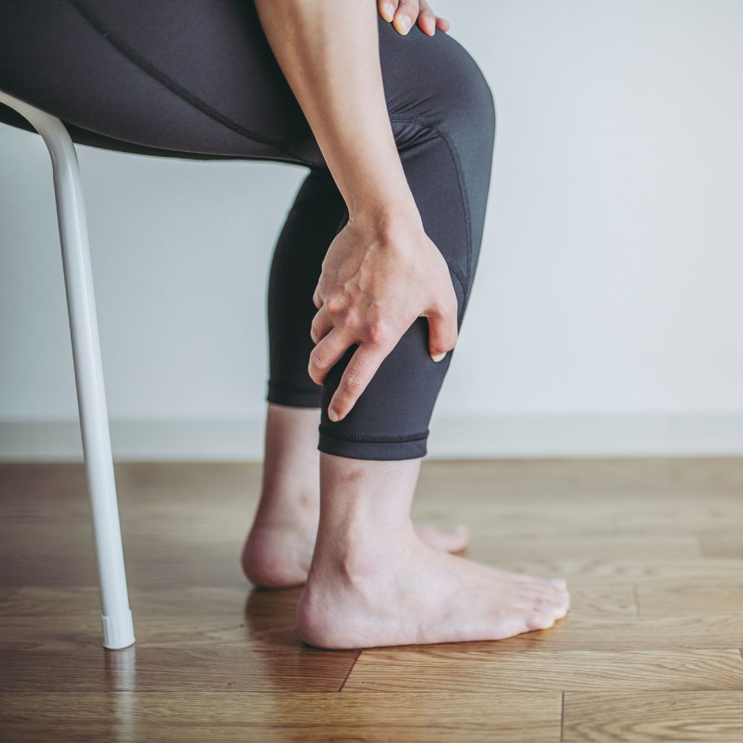 Seated person grabbing their lower calf in pain, wearing black leggings and barefoot on a wooden floor