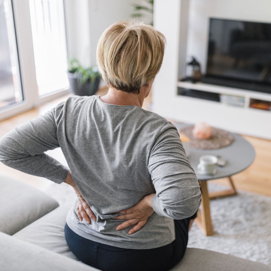 Woman sitting on a couch holding her lower back in a bright living room with a coffee table and TV