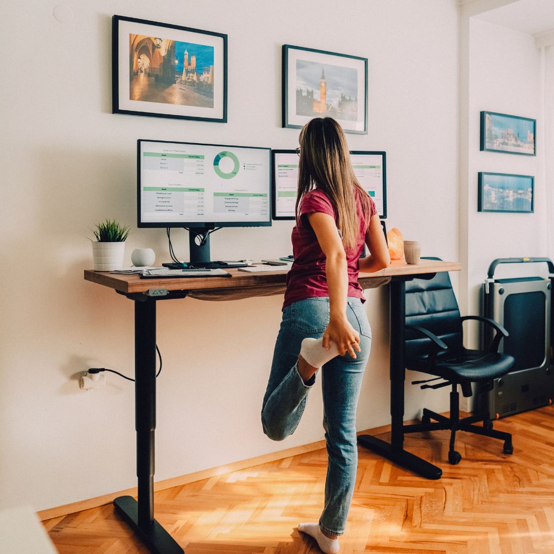 Woman stretching her quad behind her while standing at an adjustable standing desk with dual monitors in a bright home office