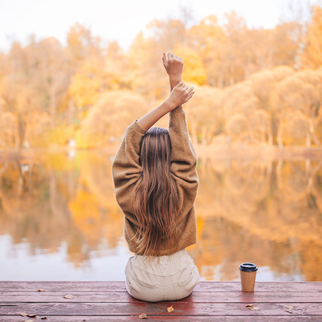 Woman sitting on a dock stretching with arms raised, facing a calm lake lined with golden autumn trees, takeaway coffee beside her