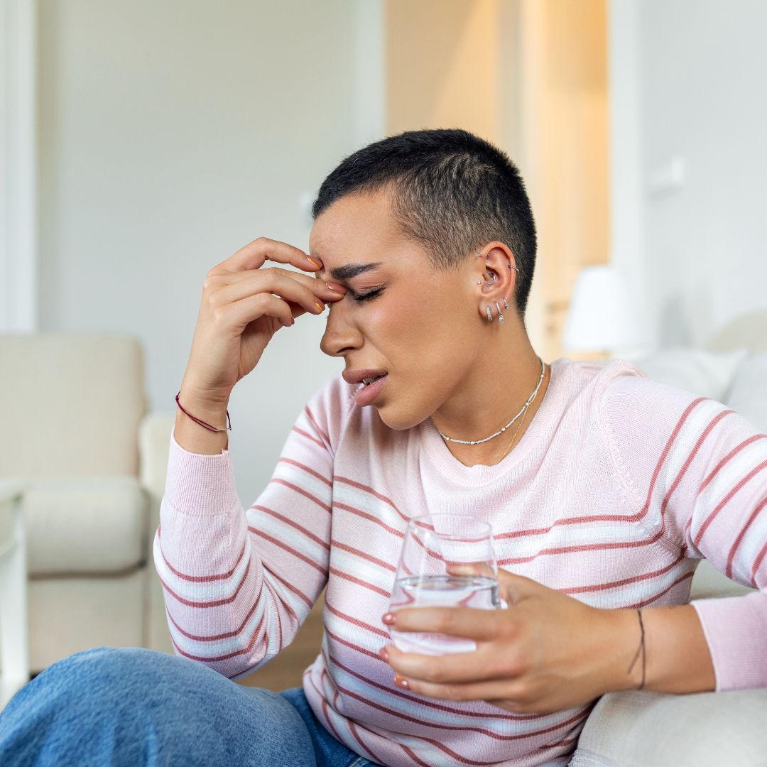 Person sitting on a couch pressing the bridge of their nose and wincing while holding a glass of water