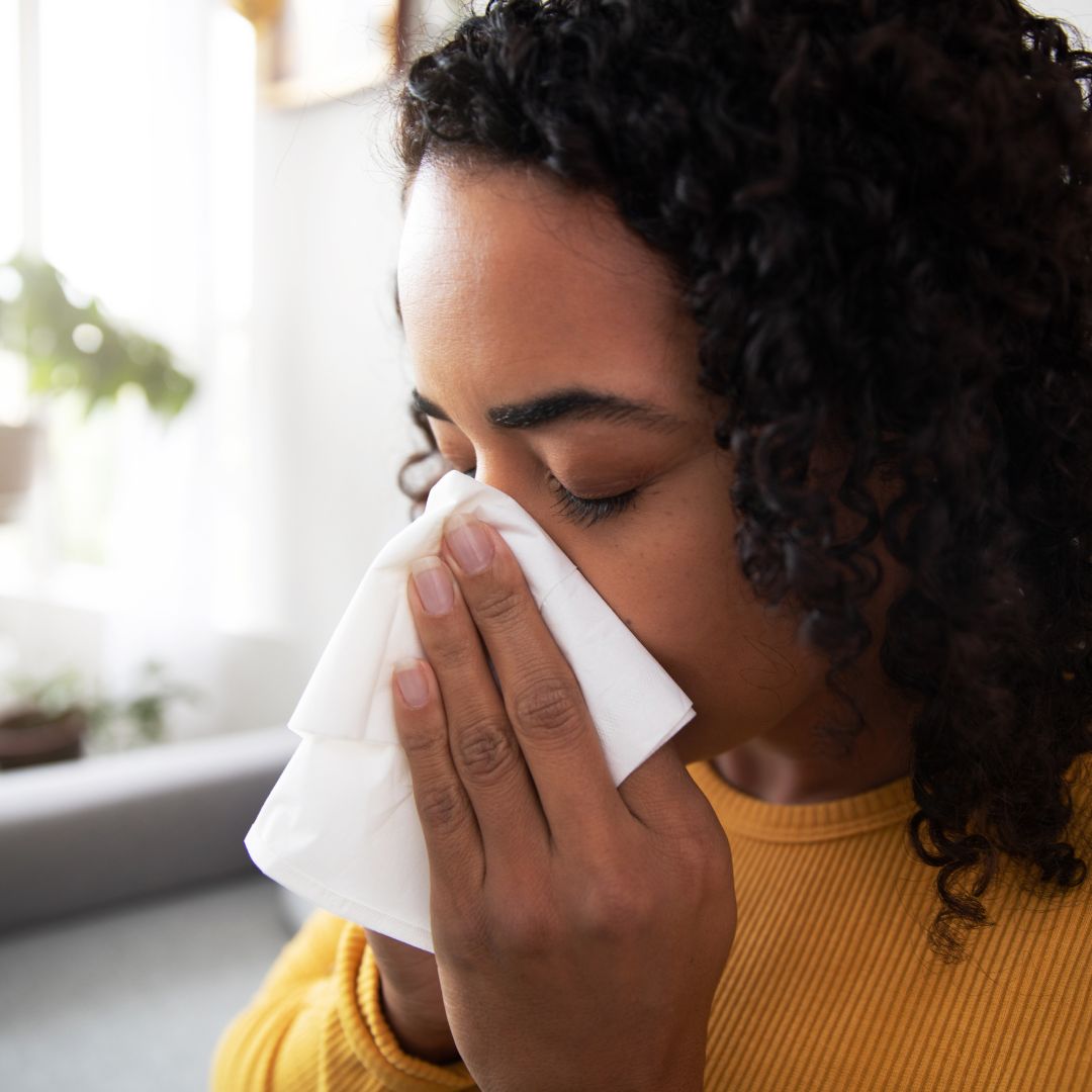 Woman blowing her nose into a tissue on a couch in a bright living room
