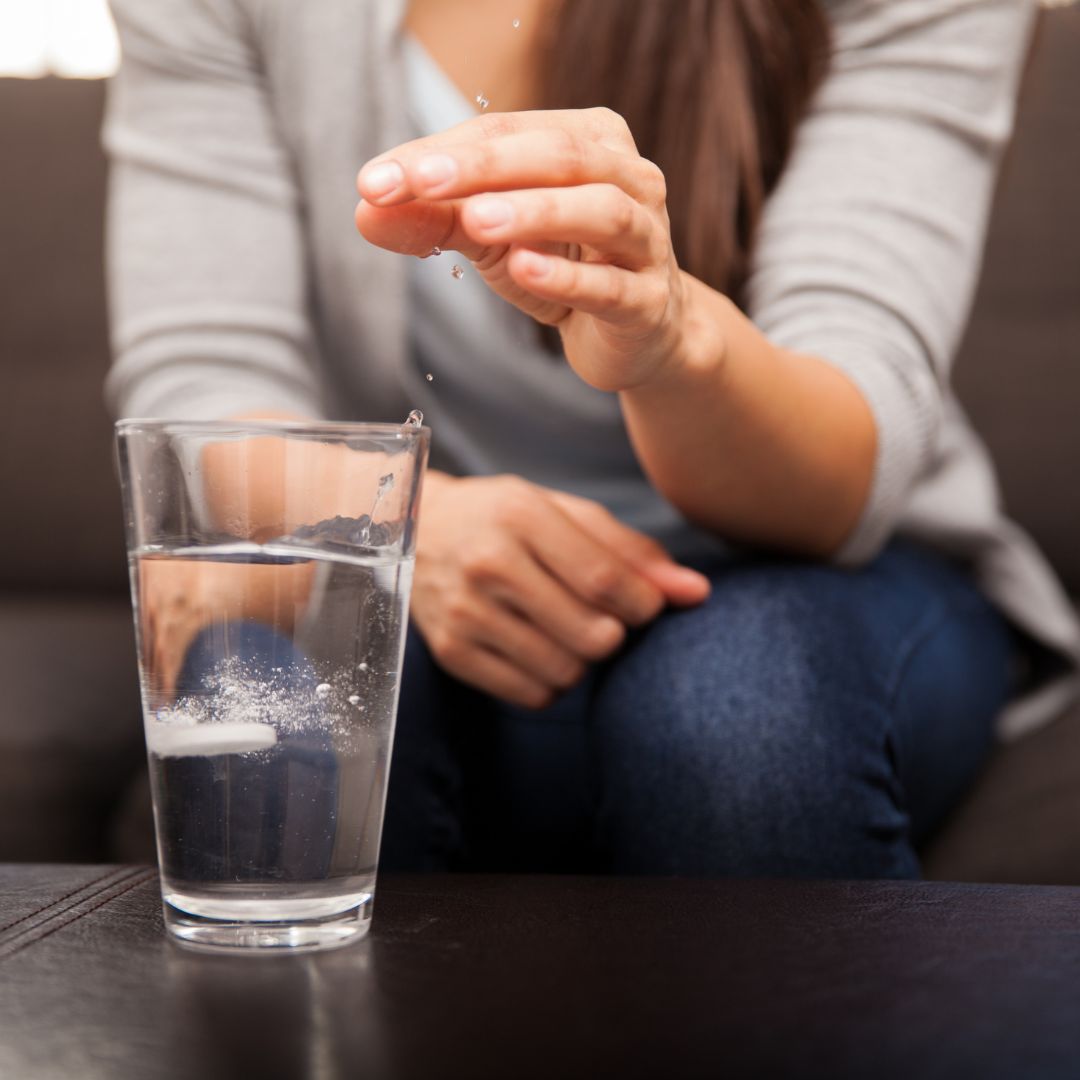 Person dropping an effervescent antacid tablet into a glass of water while seated on a couch