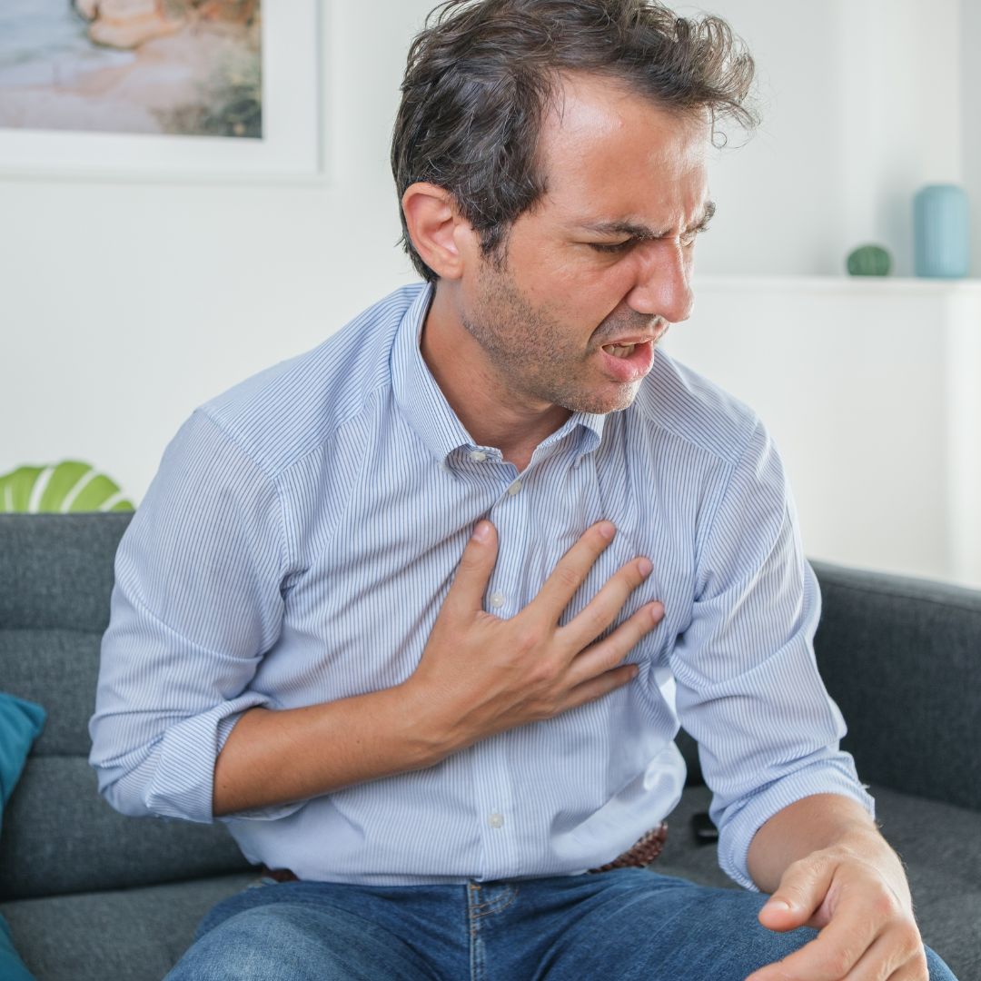 Middle-aged man clutching his chest in pain on a couch, appearing to experience heartburn or acid reflux