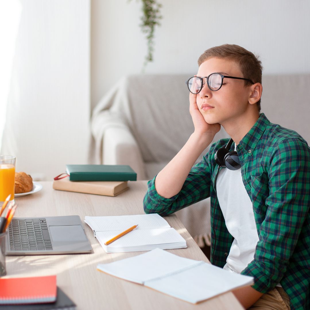 Teen boy dozing at a desk with laptop, open notebooks and headphones in a bright living room