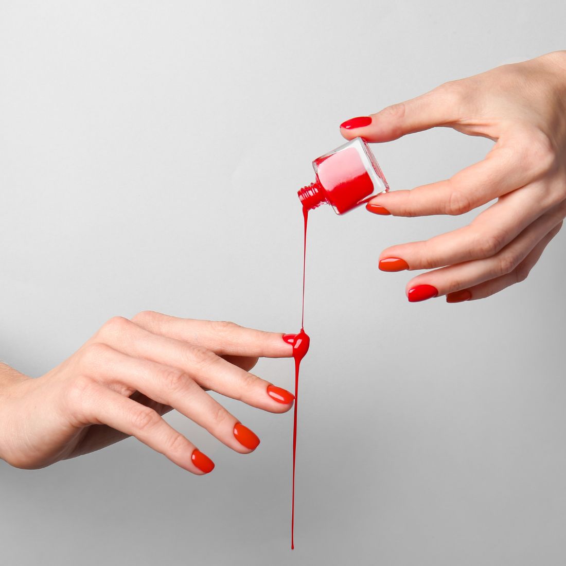 Two hands pouring red nail polish from a tipped bottle onto another finger against a plain light gray background