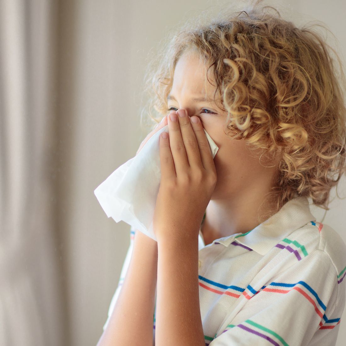 Young child sneezing into a tissue indoors, wearing a striped polo shirt