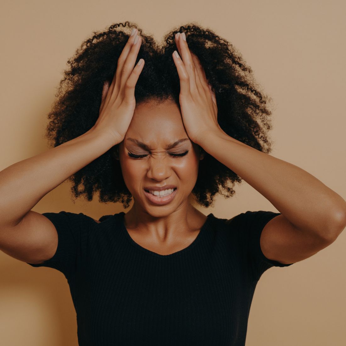 Woman clutching her head with both hands, eyes closed, wincing in pain against a beige background