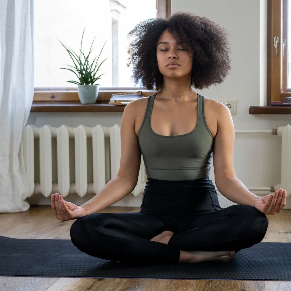 Young woman meditating cross-legged on a black yoga mat by a window, eyes closed and focusing on calm breathing
