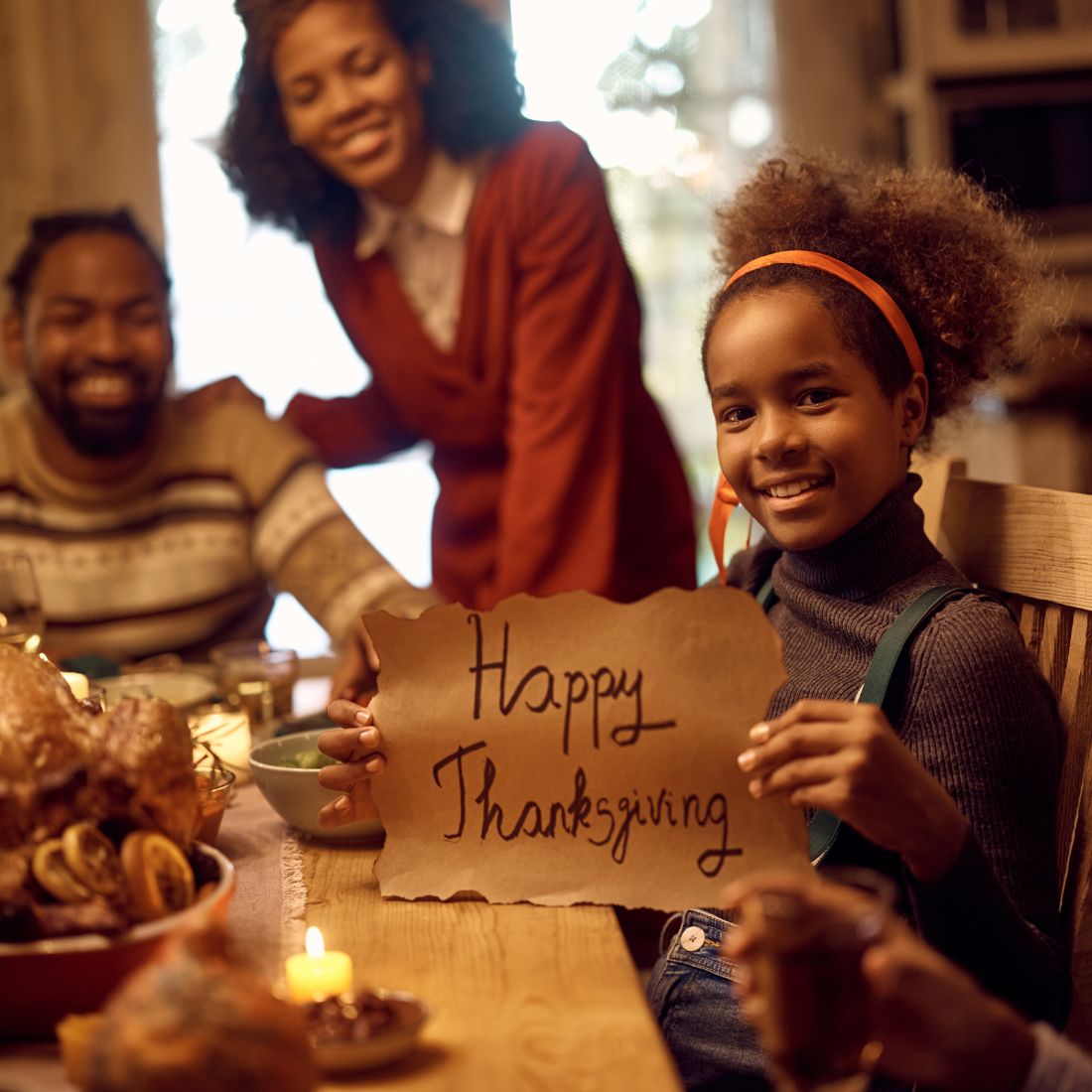 Girl smiling at a warmly lit Thanksgiving dinner table holding a handmade ‘Happy Thanksgiving’ sign with family behind her