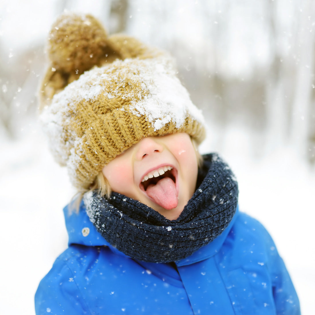 Child in blue coat and tan knit hat sticking out tongue to catch falling snow in a snowy outdoor setting
