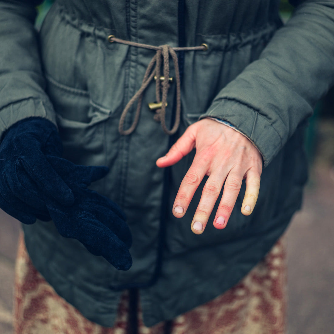 Person removing a black glove to reveal red and pale fingertips while wearing a green winter coat outdoors