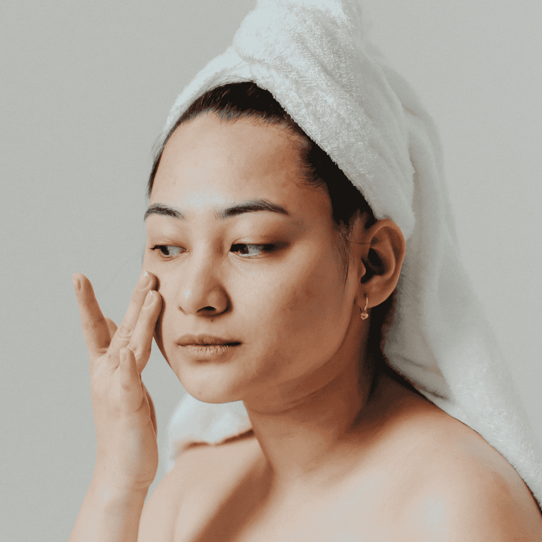 Woman with towel-wrapped hair applying moisturizer to her cheek against a plain light background