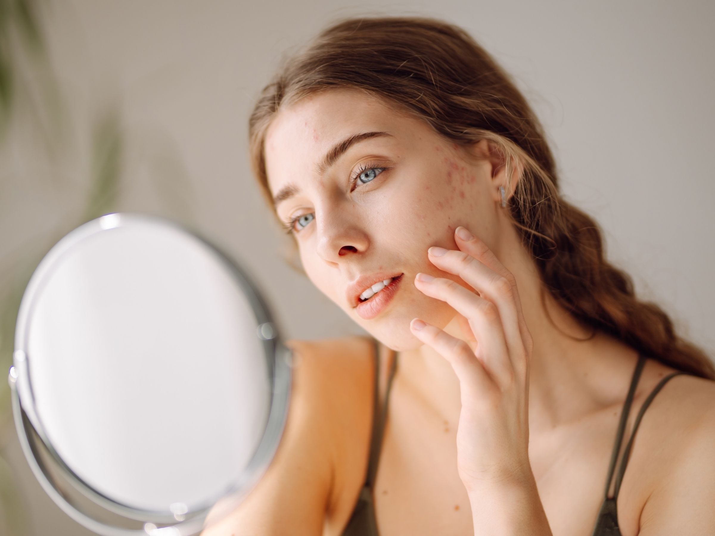 Young woman examining acne in a mirror while touching her cheek in a calm indoor setting