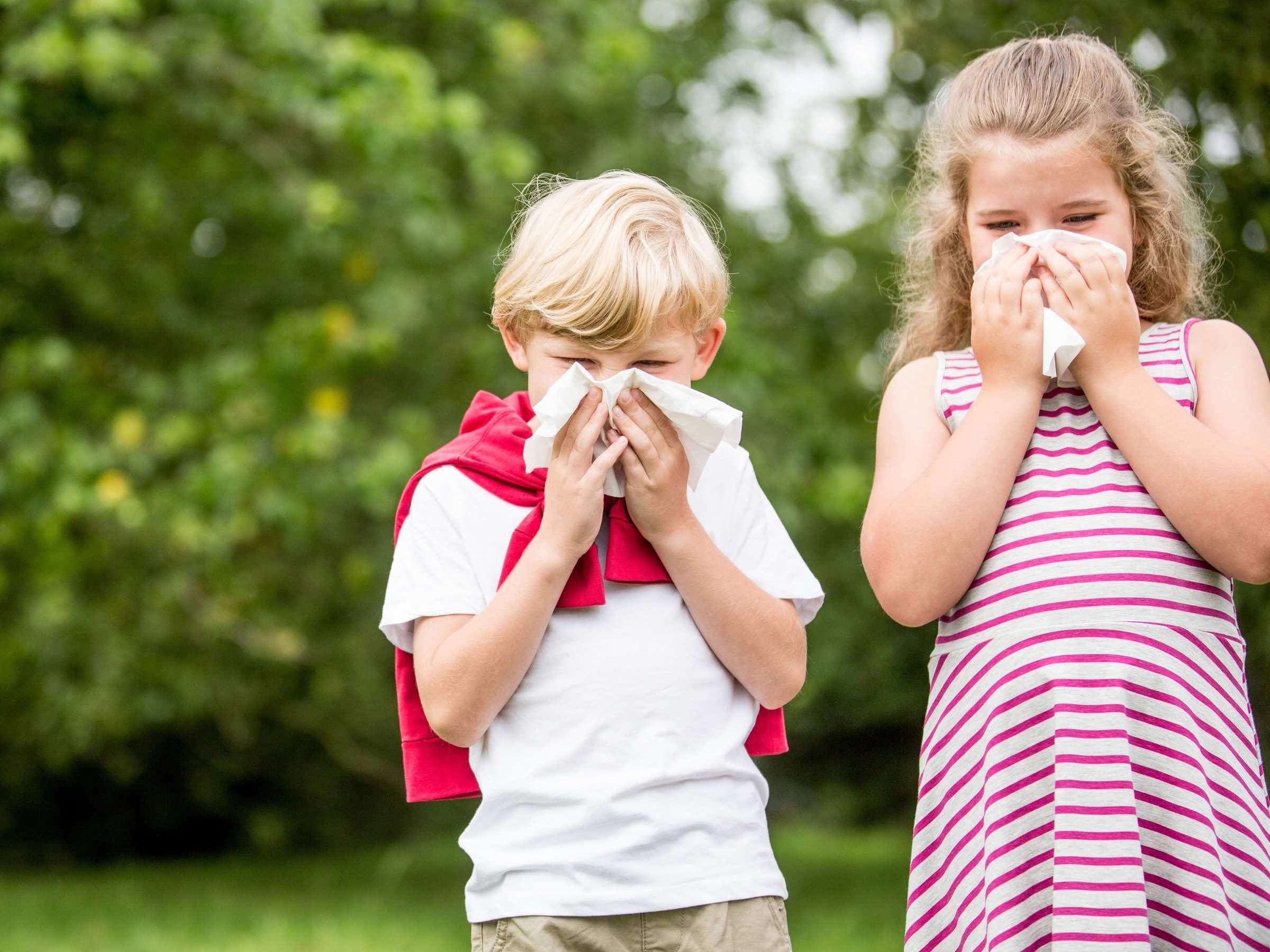 Two children outdoors in a park sneezing into tissues, illustrating seasonal allergies