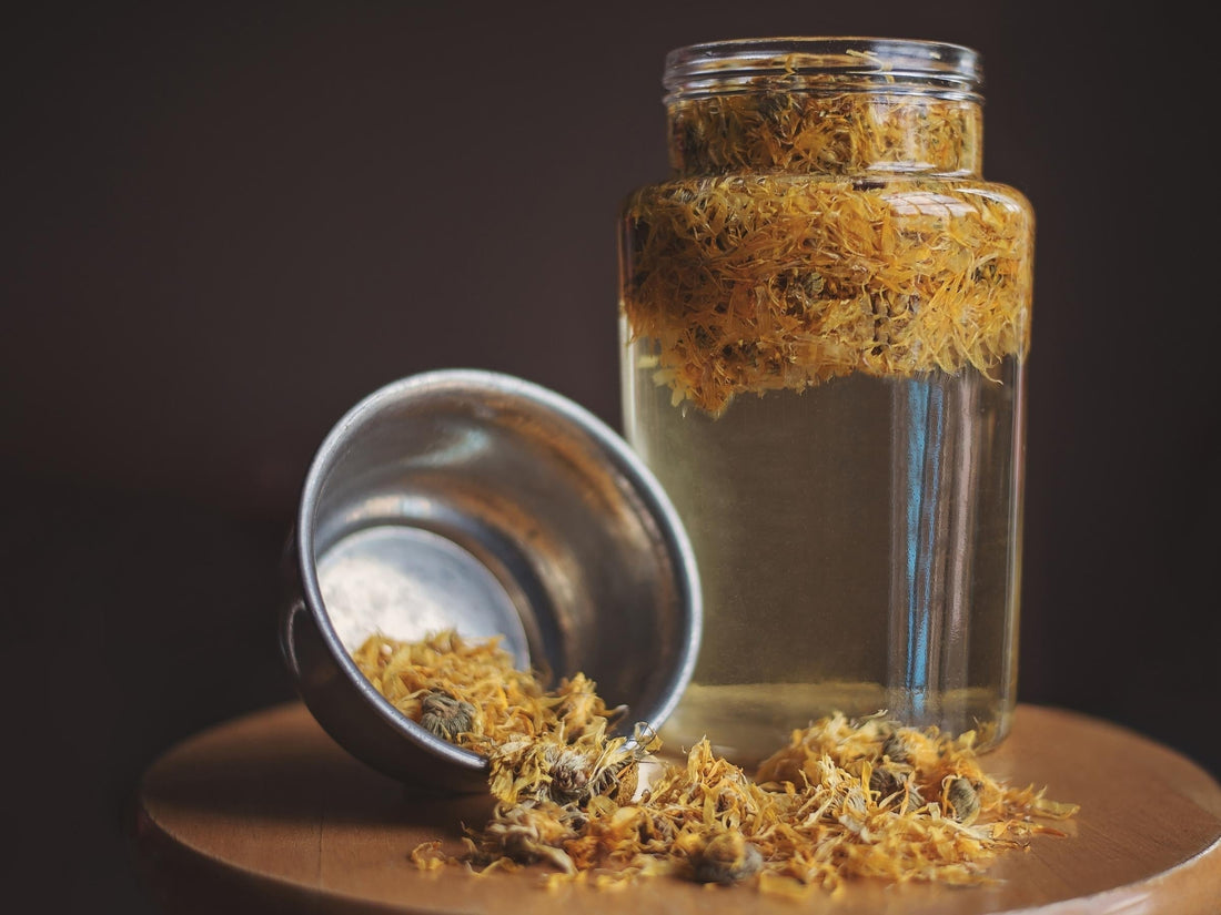 Glass jar of dried yellow flowers with a metal scoop spilling herbs onto a wooden surface, dark blurred background