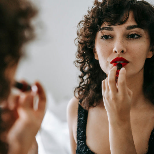 Woman with curly hair and freckles applying red lipstick in front of a mirror, symbolizing natural beauty, skincare, and lip health.