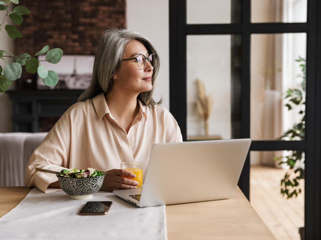 Older woman with gray hair and glasses enjoying breakfast while working on a laptop, symbolizing cognitive health, focus, and memory support.