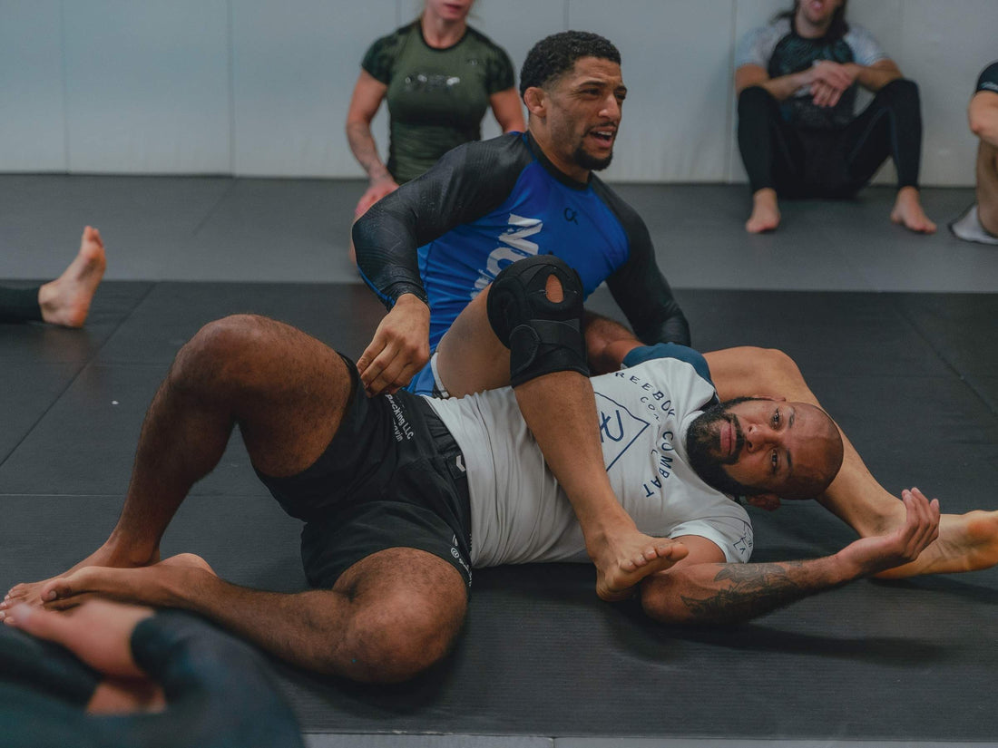 Two male athletes grappling on a black gym mat during Brazilian jiu-jitsu training, one applying a leg control while teammates watch