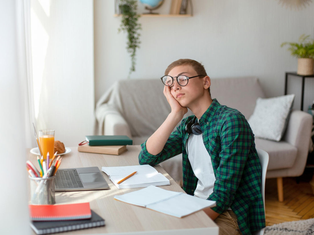 Teen boy dozing at a desk with laptop, open notebooks and headphones in a bright living room