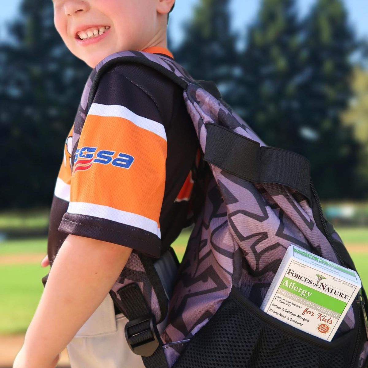 Child in orange and black jersey smiles with Forces of Nature Allergy for Kids box in his backpack.